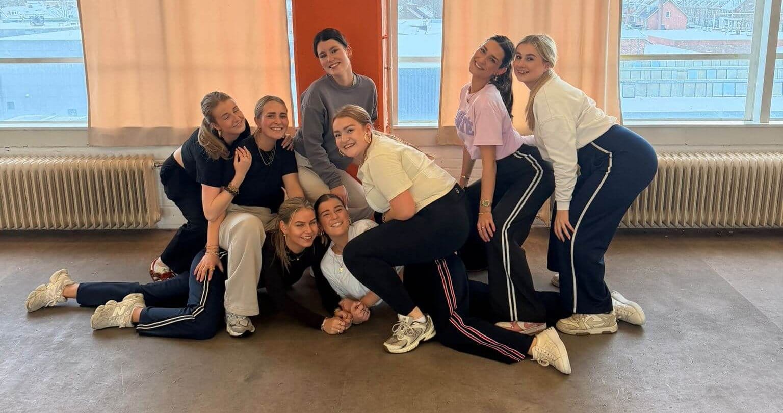 Een vrolijke groep vrouwen geniet van een creatieve chairdance-sessie tijdens een uniek bedrijfsuitje op locatie in een studio met een zwart-wit geblokte vloer.