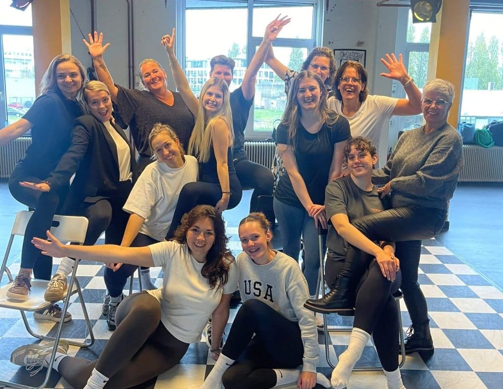Een grote, enthousiaste groep vrouwen poseert vrolijk na een actieve vriendinnen workshop in een studio met een karakteristieke geblokte vloer.